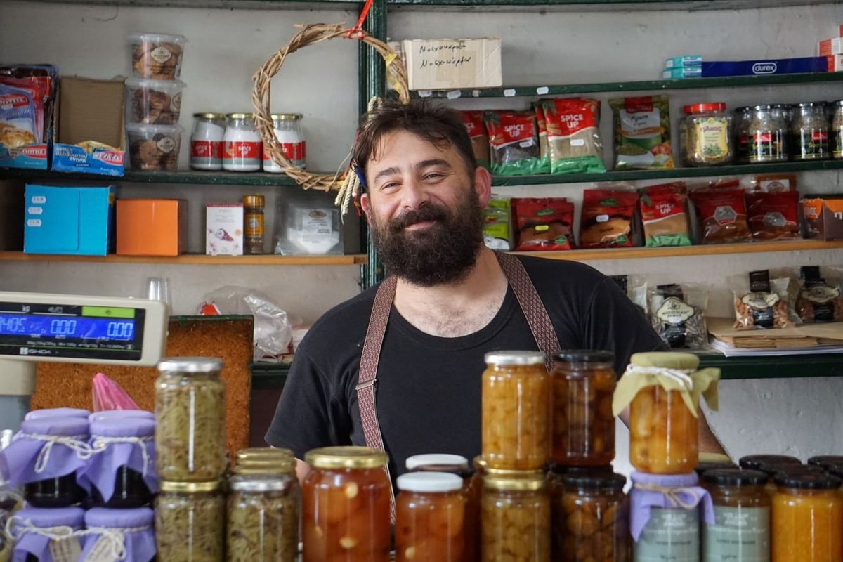 Smiling shopkeeper standing behind shelves of homemade preserves in his shop