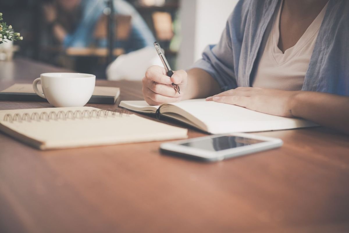 Woman writing in a notebook beside a teacup and a smartphone